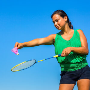 Young Colombian woman serve with badminton racket and shuttle in front of blue sky. On this sunny day in summer season the female person likes to practice sport outside. The woman is looking at the shuttle.
